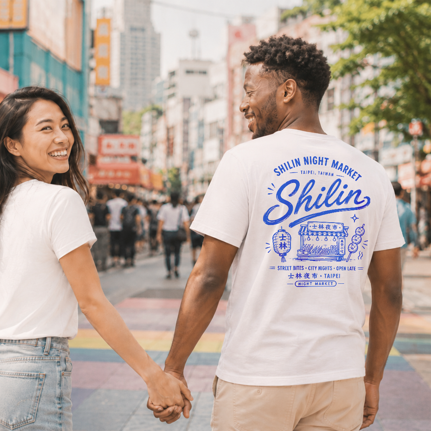Two people holding hands on a city street, both wearing white t-shirts with 'Shilin Night Market' design.